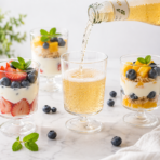 Clear plastic dessert cups filled with layered yogurt, fresh fruits, and granola, with a sparkling beverage being poured into a matching cup.